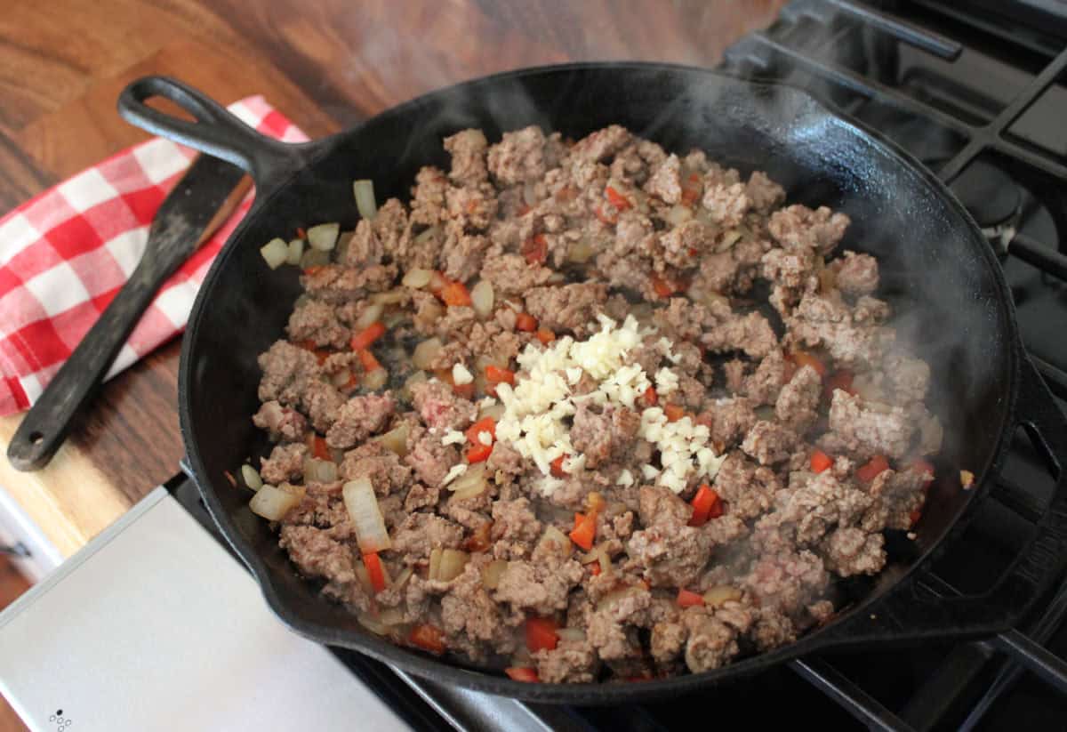 ground beef, onions, bell peppers, and garlic cooking in a cast iron skillet