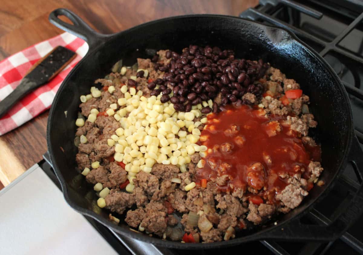 corn, black beans, and enchilada sauce being added ontop a cast iron skillet filled with cooked ground beef