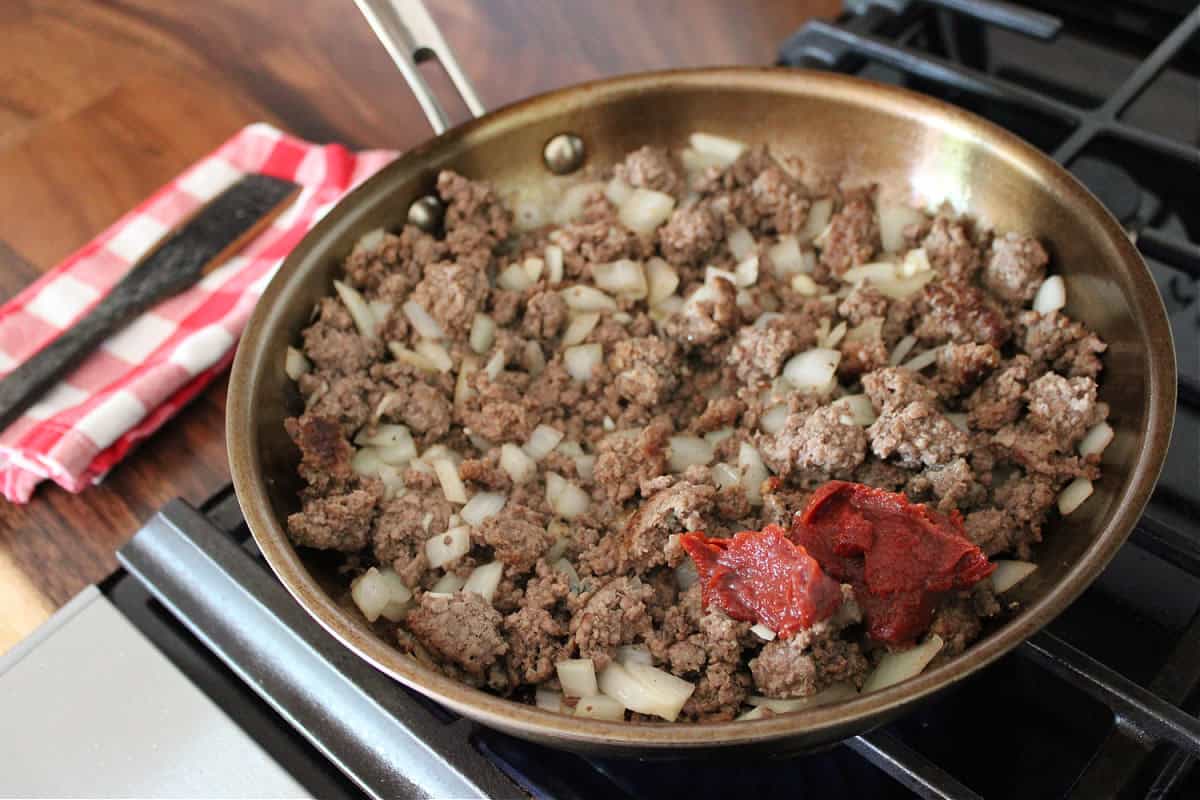 Browned ground beef and onions with a dollop of tomato paste in a pan on the stove top.