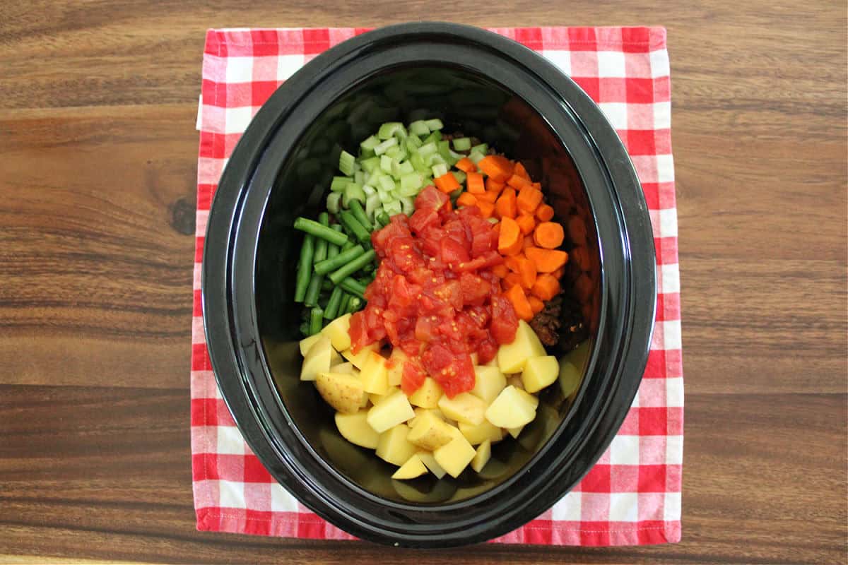 Over head shot of diced potatoes, stewed tomatoes, green beans, carrots in a black slow cooker.