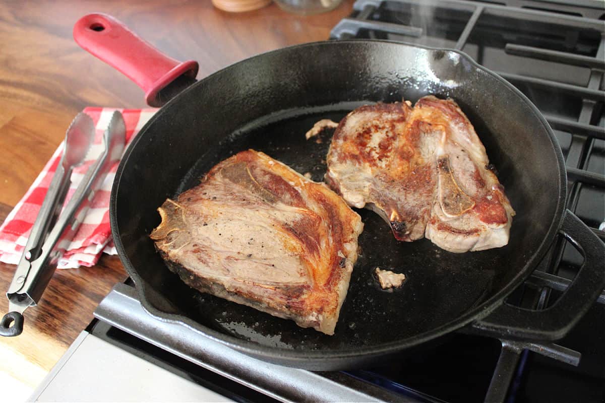 Two pork chops being browned in a black cast iron skillet.