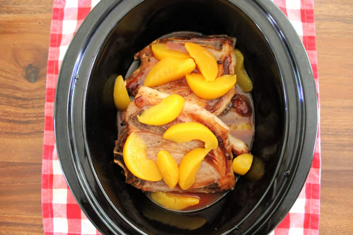 Browned pork chops and canned peaches in a black slow cooker.
